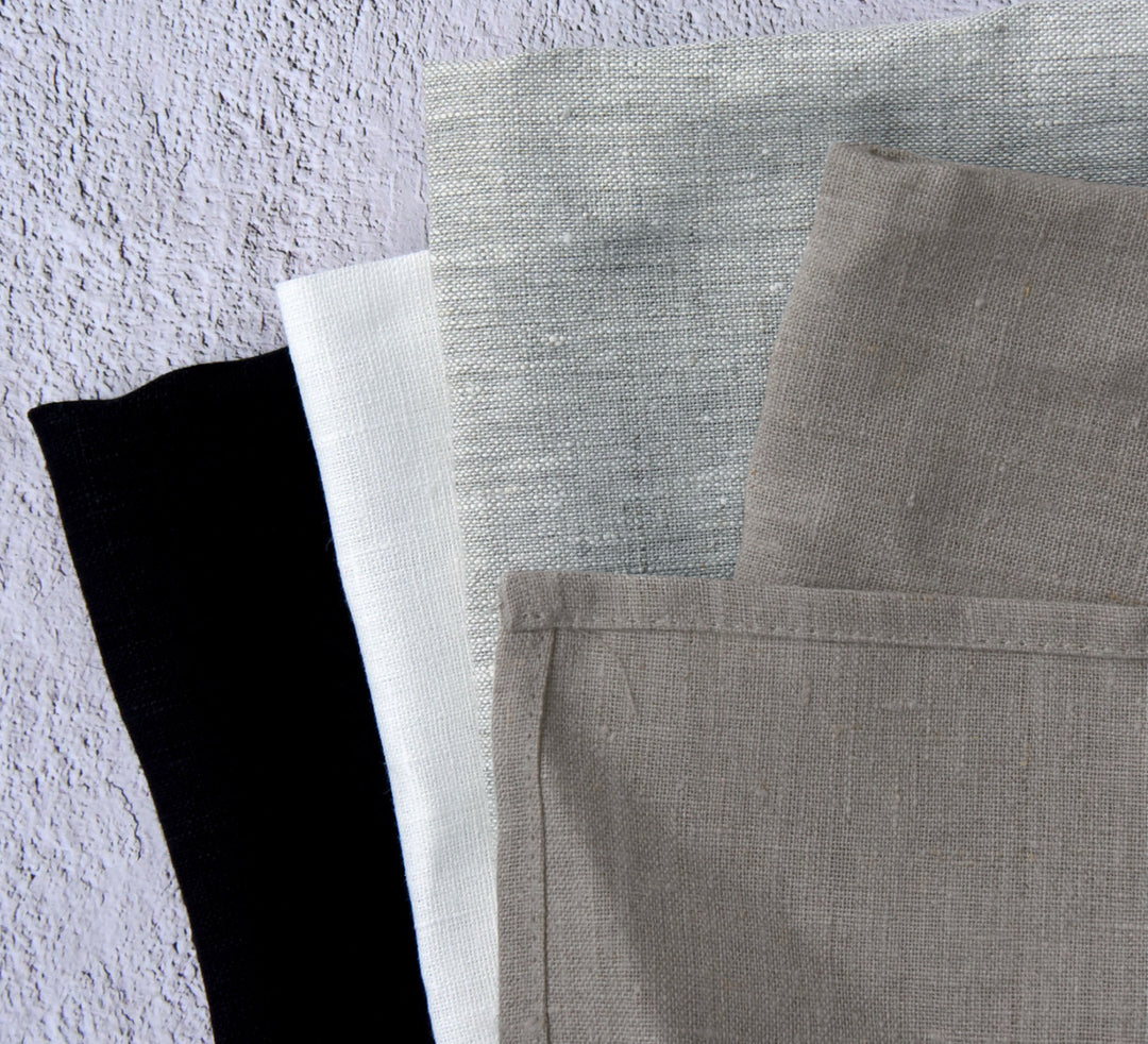 Three folded pure linen tea towels in different colors: black, white, and oatmeal, displayed against a textured background.